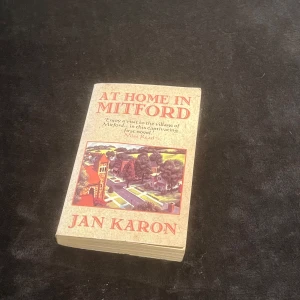 At Home in Mitford - Säljer boken 'At Home in Mitford' av Jan Karon. Boken har en beige omslag med en färgglad illustration av en by. Perfekt för dig som gillar mysiga och hjärtevärmande berättelser. Boken är i pocketformat och lätt att ta med sig. Perfekt för en lugn läsning hemma eller på resan.