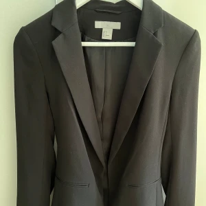 Svart kavaj från H&M - Säljer en stilren svart kavaj från H&M i storlek 34. Den har långa ärmar och ett bälte i midjan för en snygg passform. Perfekt för både vardag och fest!