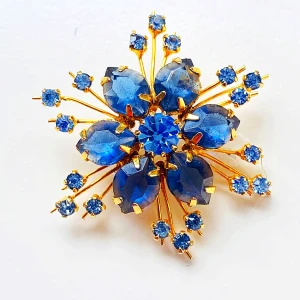Blå och guld brosch med blommotiv - Säljer en superfin vintage brosch i form av en blomma med gnistrande blå stenar och en elegant guldfärgad bas. Perfekt för att piffa upp vilken outfit som helst! Den är ungefär 5 cm i diameter och har en säkerhetsnål på baksidan för enkel fastsättning.