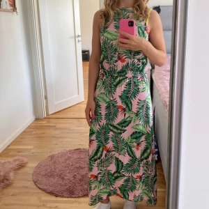 Tropisk långklänning med bladmönster - Säljer en superfin långklänning med tropiskt bladmönster i grönt och rosa. Klänningen är ärmlös och har en avslappnad passform, perfekt för sommarens fester eller en dag på stranden. Materialet känns lätt och luftigt, vilket gör den idealisk för varma dagar. 🌿🌸