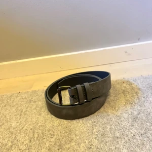Brunt bälte i mocka - Säljer ett snyggt brunt/grå bälte i mocka. Det har en stilren design med en enkel spänne i metall. Perfekt för att ge en extra touch till din outfit. Passar både till vardags och fest!