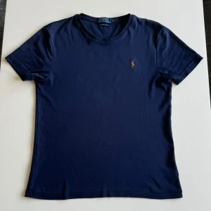 Mörkblå t-shirt från Ralph Lauren - Säljer en snygg mörkblå t-shirt från Ralph Lauren, Custom Slim Fit-modellen. Den är perfekt för både vardag och när du ska ut på något kul. Måtten är 63 cm lång (från högsta punkt) och 47 cm bred (från armhåla till armhåla), så den sitter skönt men lite slimmad. Skicket är 8/10, inga trasigheter eller sånt, bara använd några gånger. Nypriset är runt 1000-lappen och säljer den för 320kr. Lämna ett dm om du har några frågor eller funderingar!