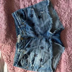 Blå jeansshorts med stjärnor - Säljer ett par coola blå jeansshorts med slitna kanter och broderade stjärnor. Perfekta för sommaren och ger en avslappnad look. De har en klassisk femficksdesign och knappar framtill. Passar perfekt för en dag på stranden eller en somrig festival!