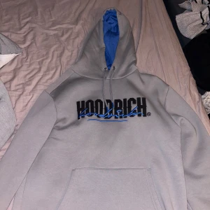 Grå hoodie från Hoodrich - Säljer en snygg grå hoodie från Hoodrich med blå detaljer. Den har en stor logga på både fram- och baksidan. Insidan av huvan är blå. Perfekt för en avslappnad stil! Säljer för 500 