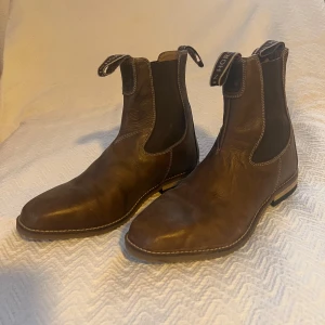 Bruna Chelsea boots i skinn - Säljer ett par snygga bruna Chelsea boots i skinn. De har en klassisk design med elastiska paneler på sidorna och en dragflik baktill för enkel påtagning. Perfekta för både höst och vinter, och de ger en stilren look till vilken outfit som helst.