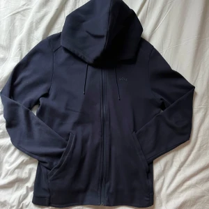 Marinblå hoodie från Boss - Säljer en stilren marinblå hoodie från Boss med dragkedja och huva. Perfekt för en avslappnad look. Den har en diskret logga på bröstet och praktiska fickor framtill. Nypris 1799:- SEK 