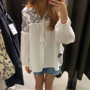 Vit blus med broderade detaljer - Säljer en luftig vit blus med vackra blå broderade detaljer vid halsen. Blusen har långa ärmar och en knytning framtill som ger en bohemisk känsla. Perfekt till sommaren. Säljer för att den inte kommer till användning 