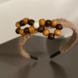 Beige diadem med träpärlor - Snyggt beige diadem dekorerat med träpärlor i brunt, vitt och orange. Perfekt accessoar för att ge en naturlig touch till din stil. Diademet har en flätad design som ger en bohemisk känsla.