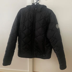 Svart quiltad jacka från Vinson Polo Club - Säljer en svart quiltad jacka från Vinson Polo Club. Jackan har en dragkedja framtill och en huva för extra skydd. Den är perfekt för kyligare dagar och har ett stilrent märke på ärmen. Passar bra för en casual look.