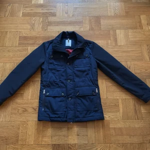 Mörkblå fieldjacka med fodring - Snygg mörkblå jacka med knappar och dragkedja. Jackan har flera fickor och en stilren design. Perfekt för kyligare dagar med sin långa ärmar och praktiska detaljer.