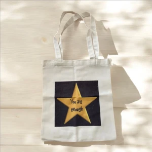 Vit shoppingväska med stjärna - Snygg vit shoppingväska med ett tryck av en gul stjärna och texten 'You are enough'. Perfekt för att bära med sig det nödvändigaste med stil. Väskan har två handtag och är tillverkad i ett slitstarkt material.