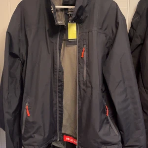 Svart vindjacka från Helly Hansen - Snygg marinblå vindjacka från Helly Hansen med dragkedjor i kontrasterande röd färg. Jackan har en hög krage och praktiska fickor med dragkedja. Perfekt för blåsiga dagar och har en sportig look.