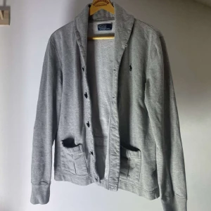 Grå cardigan från Polo Ralph Lauren - Säljer en stilren grå cardigan från Polo Ralph Lauren. Den har långa ärmar och knappar framtill. Perfekt för en avslappnad look. Den ikoniska loggan är broderad på bröstet. Passar bra till både jeans och chinos.