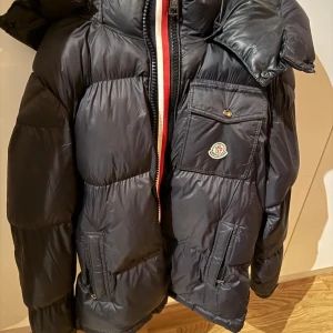 Svart dunjacka från Moncler - Säljer en snygg svart dunjacka från Moncler med hög krage och huva. Jackan har en dragkedja framtill och en bröstficka med knapp. Perfekt för kalla dagar!