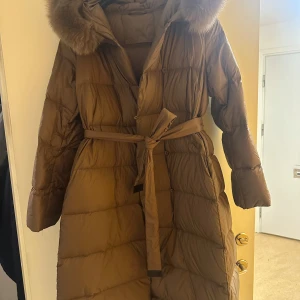 Brun dunjacka med pälsdetalj - Säljer en lång brun dunjacka med pälsdetalj på huvan. Jackan har ett bälte i midjan och knappar framtill. Perfekt för kalla dagar med sin varma och mysiga design.
