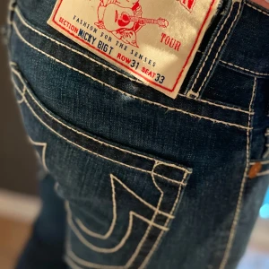 Mörkblå true religion jeans.  Mickey Big T - Snygga mörkblå jeans med vita kontrastsömmar och unika bakfickor. Jeansen har en klassisk femficksdesign och en bekväm passform. Perfekta för en stilren look.. aningen vida jeans för de som gillar de.. Köpt i USA. unika eftersom bakfickorna saknar flärpen. Jag är 1,83 lång. Gammal  hockeyspelare så jag har svårt att få på dom över låren. Väldigt svåra att få tag på. Knappt använda 31/33 enligt usa men passar mig som brukar ha 34 normalt i midja. . Skulle säga att dom är som en 32-33 i midjan,