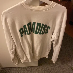 Vit sweatshirt med texten 'Paradise' - Säljer en vit sweatshirt med texten 'Paradise' i grönt på framsidan. Tröjan har långa ärmar och en ribbad krage. Perfekt för en avslappnad stil.