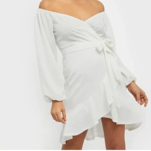 Studentklänning/ vit klänning - Säljer en elegant vit offshoulder klänning med omlott-design och knytband i midjan. Klänningen har långa ärmar och en volangdetalj vid fållen. Perfekt för studenten, midsommar, eller bara sommaren! Oanvänd