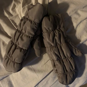 Grå/beiga vadderade tumvantar - Säljer ett par grå/beiga vadderade tumvantar som är perfekta för kalla dagar. De har en quiltad design med elastiska muddar för extra värme och komfort. Passar bra för vinteraktiviteter och håller händerna varma och sköna. Helt nya med prislapp kvar.