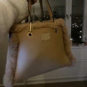 Beige handväska med pälsdetaljer - Snygg beige handväska med mjuka pälsdetaljer längs kanterna. Väskan har två handtag och en elegant metallplatta framtill. Perfekt för att ge din outfit en lyxig touch.