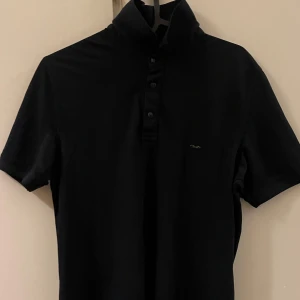 Svart kortärmad t-shirt från Jeuss Exclusive - Snygg svart t-shirt från Jeuss Exclusive med korta ärmar och knappar vid halsen. Perfekt för en stilren look. Passar bra till både jeans och chinos.