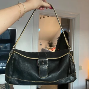 Svart axelväska med spänne❤️ - Snygg svart axelväska i skinn med kontrasterande sömmar och ett stilrent spänne framtill. Väskan har en praktisk storlek och en justerbar axelrem. Perfekt för att bära dina dagliga nödvändigheter med stil.