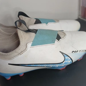 Nike Air Zoom fotbollsskor - Snygga Nike Air Zoom fotbollsskor i vitt med blå och svarta detaljer. Skorna har en unik design med blå sula och rosa dobbar, samt ett elastiskt band över vristen för extra stöd. Perfekta för fotbollsplanen med sin moderna look och funktionalitet.