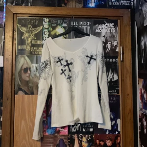 Vit långärmad tröja med korsdetaljer - Säljer en vit långärmad tröja med coola korsdetaljer och vingmönster. Tröjan har en v-ringad hals och är perfekt för en unik stil. Passar bra till både jeans och kjol. Hör av dig vid intresse!