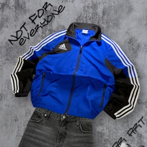 Blå och svart vindjacka från Adidas - Säljer en blå och svart vindjacka från Adidas med klassiska vita ränder längs ärmarna. Jackan har dragkedja och Adidas-logga på bröstet. Perfekt för en sportig look! Passar M & L, främst M