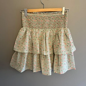 Grön blommig kjol med volanger - Charmig grön kjol med blommigt mönster och volanger. Den har en elastisk midja för bekväm passform. Perfekt för en somrig look!