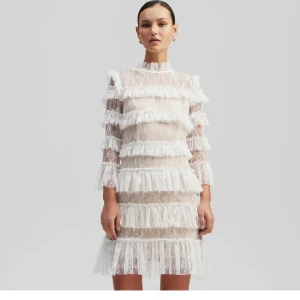 Vit spetsklänning med volanger - Elegant vit spetsklänning med volanger och långa ärmar. Klänningen har en transparent design med flera lager av spets och volanger som ger en romantisk känsla. Perfekt för speciella tillfällen.