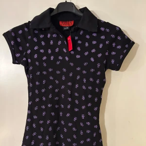 Svart kortärmad polotopp med lila dödskallemönster - Snygg svart polotopp från Poizen Industries med lila dödskallemönster. Toppen har korta ärmar och en klassisk krage. Perfekt för en unik och edgy stil.