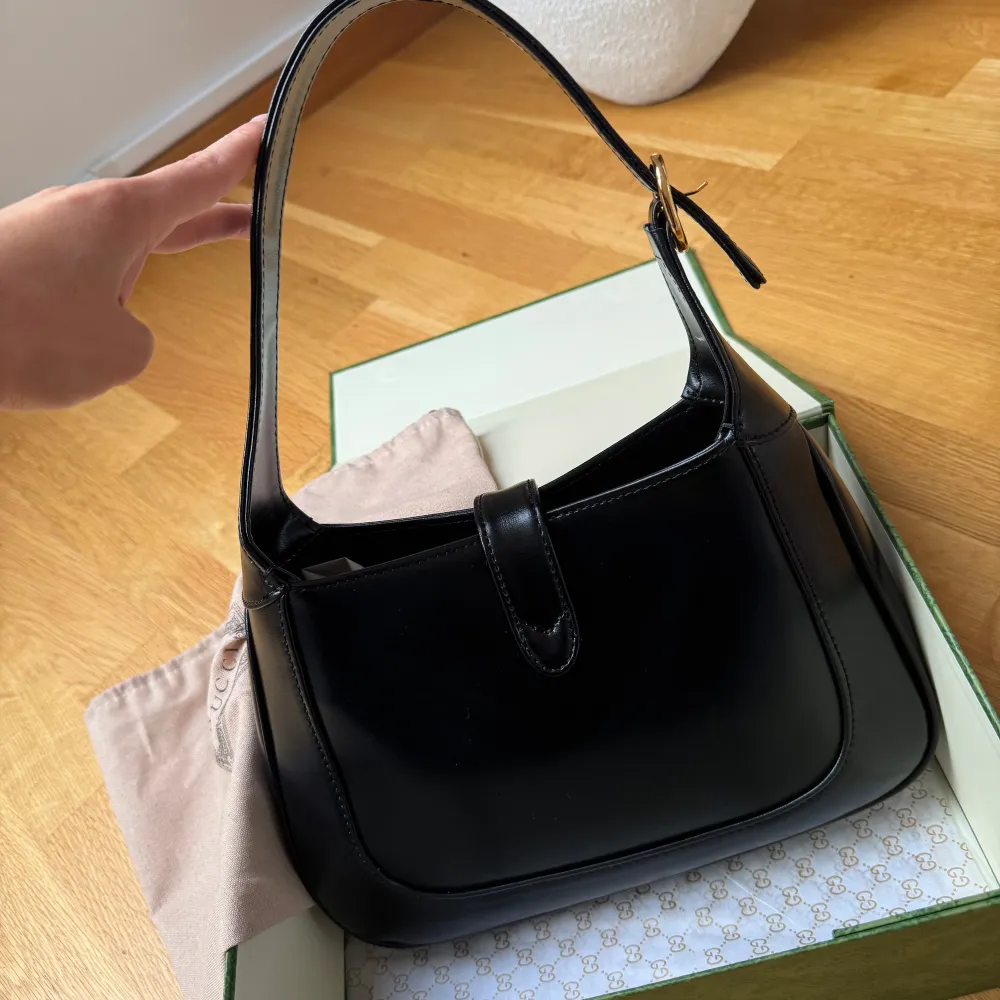 Elegant svart axelväska i skinn med stilrena gulddetaljer. Väskan har en justerbar axelrem och en dekorativ spänne framtill. Perfekt för att ge en lyxig touch till din outfit. Box och dustbag ingår!. Laukut & Käsilaukut.