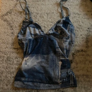 Jeansinspirerad korsettopp - Snygg jeansinspirerad korsettopp med justerbara axelband och dekorativt spänne. Toppen har en unik tvättad look med slitna detaljer och en figurnära passform. Perfekt för en trendig och avslappnad stil.