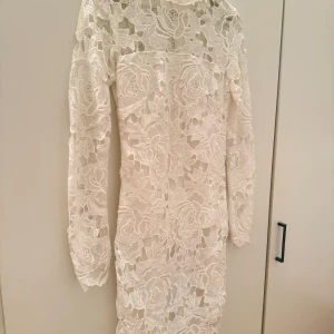 STUDENTKLÄNNING  - Elegant vit spetsklänning med långärmad design och vackert blommönster. Perfekt för speciella tillfällen med sin sofistikerade look. Klänningen har en kroppsnära passform som framhäver figuren. Skicka pm för bild på.