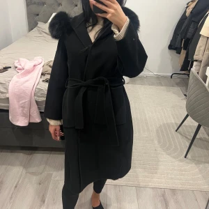 Svart kappa med pälsdetaljer - Snygg svart kappa med stor huva och pälsdetaljer. Kappan har ett knytbälte i midjan och långa ärmar. Perfekt för kyliga dagar.