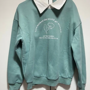 Grön sweatshirt med vit krage - Säljer en stilren grön sweatshirt med vit krage och tryck framtill. Texten 'love makes the world go around' och en blomma ger en unik touch. Perfekt för en avslappnad stil. Jätte bekväm och är som ny