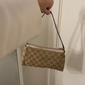 Beige axelväska från Gucci - Snygg beige axelväska från Gucci med det klassiska GG-mönstret. Väskan har en smidig dragkedja och en elegant axelrem. Perfekt för att piffa upp en outfit. Köpt och äkthetskontrollerad på Vestiaire för 4500kr, öppen för prisförslag. 