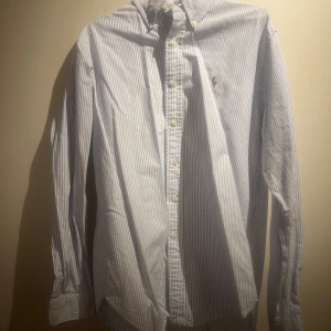 Randig skjorta från Ralph Lauren - Snygg och klassisk långärmad skjorta från Ralph Lauren i blå och vit randigt mönster. Skjortan har knappar framtill och en broderad logotyp på bröstet. Perfekt för en stilren look. Lite skrynklig efter som har legat i garderoben 