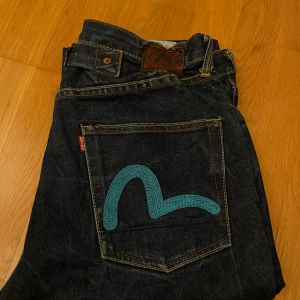 Mörkblå jeans med broderad bakficka - Snygga mörkblå evisu  jeans med en unik turkos broderad detalj på bakfickorna. Klassisk femficksdesign med gul kontrastsöm. Perfekta för en avslappnad stil. Den förra ägaren klippte upp byxorna där nere så att de skulle falla över skorna. 