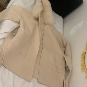 Beige kappa med huva - Säljer en stilren beige kappa med huva och pälsdetaljer. Jackan har långa ärmar och praktiska fickor framtill. Perfekt för kyligare dagar och ger en elegant touch till din outfit. Från vivanilla och original pris: 3799. Helt oanvänd då den är för stor på mig.