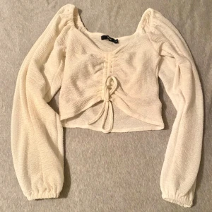 Vit långärmad topp med knyt - Säljer en söt vit topp med långa ärmar och knytning framtill. Toppen har en lätt textur och är perfekt för både vardag och fest. Den har en croppad passform och är gjord i ett mjukt material.