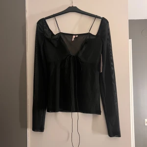 Svart långärmad topp från Nelly - Snygg svart långärmad topp från Nelly med smala axelband och knytning framtill. Perfekt för en stilren look. Toppen har en elegant design som passar till många tillfällen.