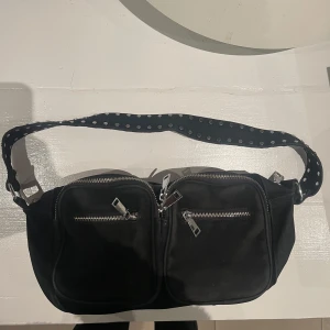 Svart axelväska från Noella - Snygg svart axelväska från Noella med två dragkedjefickor framtill. Väskan har en axelrem med nitar. 