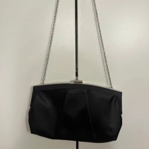 Svart kuvertväska med kedja - Elegant svart kuvertväska med en stilren kedja som axelrem. Väskan har en metallram och ett enkelt lås upptill. Perfekt för en kväll ute eller en speciell tillställning.