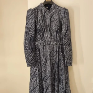 Mönstrad långklänning i glitter  - Elegant långklänning med ett fint glans. Klänningen har långa ärmar och en hög krage som ger en sofistikerad look. Perfekt för speciella tillfällen där du vill sticka ut. Materialet är av hög kvalitet och ger en fin fall.