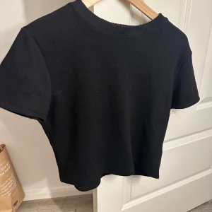 Svart kortärmad topp från Zara - Säljer en stilren svart topp från Zara. Den är kortärmad och har en rund halsringning. Perfekt för en minimalistisk look. Den är använd lappen håller på att lossna och för information är den i en kortare modell! Storleken är L men den är liten  