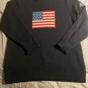 Svart stickad tröja från Gant - Säljer en svart stickad tröja från Gant med ett broderat amerikanskt flaggmotiv på framsidan. Tröjan har långa ärmar och ribbade muddar. Perfekt för en stilren och avslappnad look.