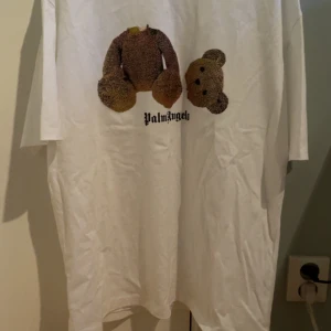 Vit Palm Angels T-shirt - Säljer en vit t-shirt från Palm Angels med ett unikt nalletryck på framsidan och Palm Angels-logga på baksidan. T-shirten har en avslappnad passform och korta ärmar. Perfekt för en cool och avslappnad stil.