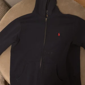 Svart hoodie från Ralph Lauren - Säljer en stilren svart hoodie från Ralph Lauren med dragkedja och den klassiska röda loggan på bröstet. Perfekt för en avslappnad look. Den har en praktisk huva och fickor framtill.